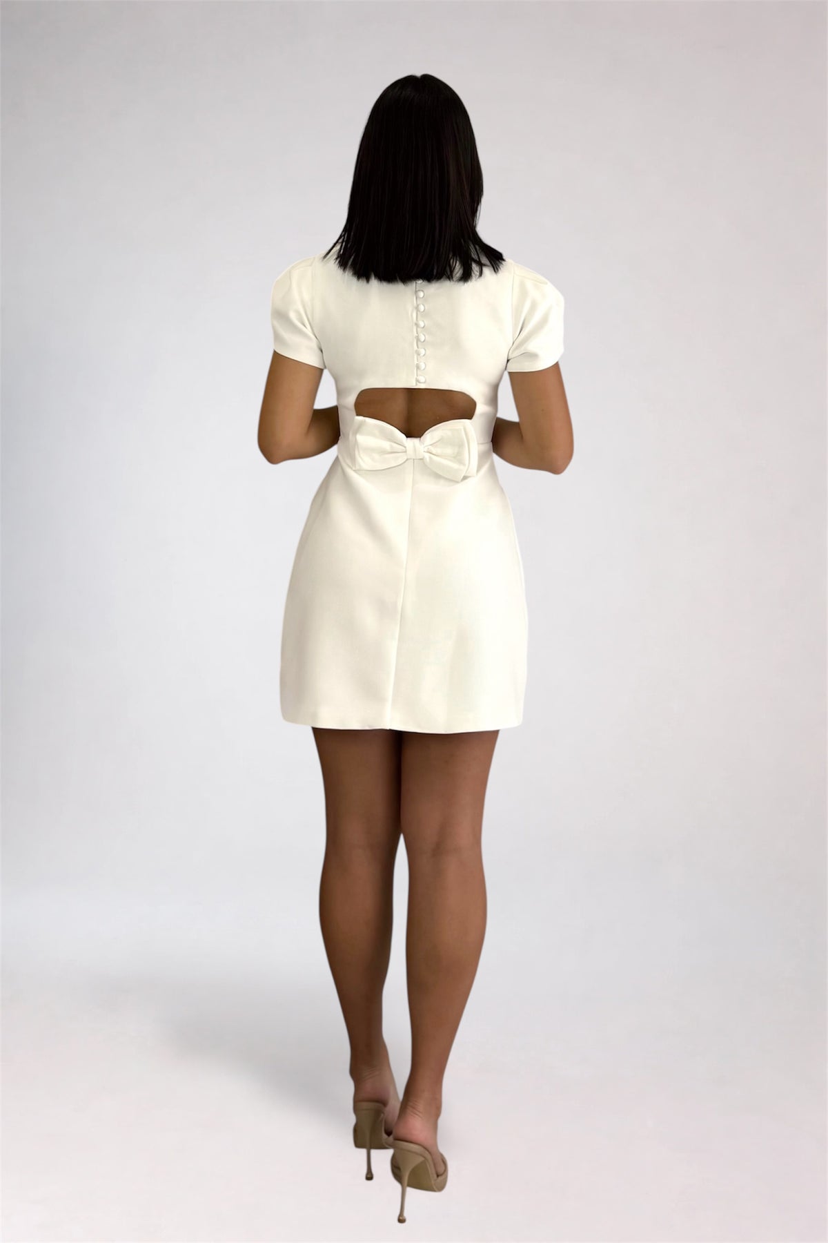 A woman viewed from behind wearing a short cream-colored dress with a cutout at the back and a bow detail, paired with beige high heels, against a light gray studio background.