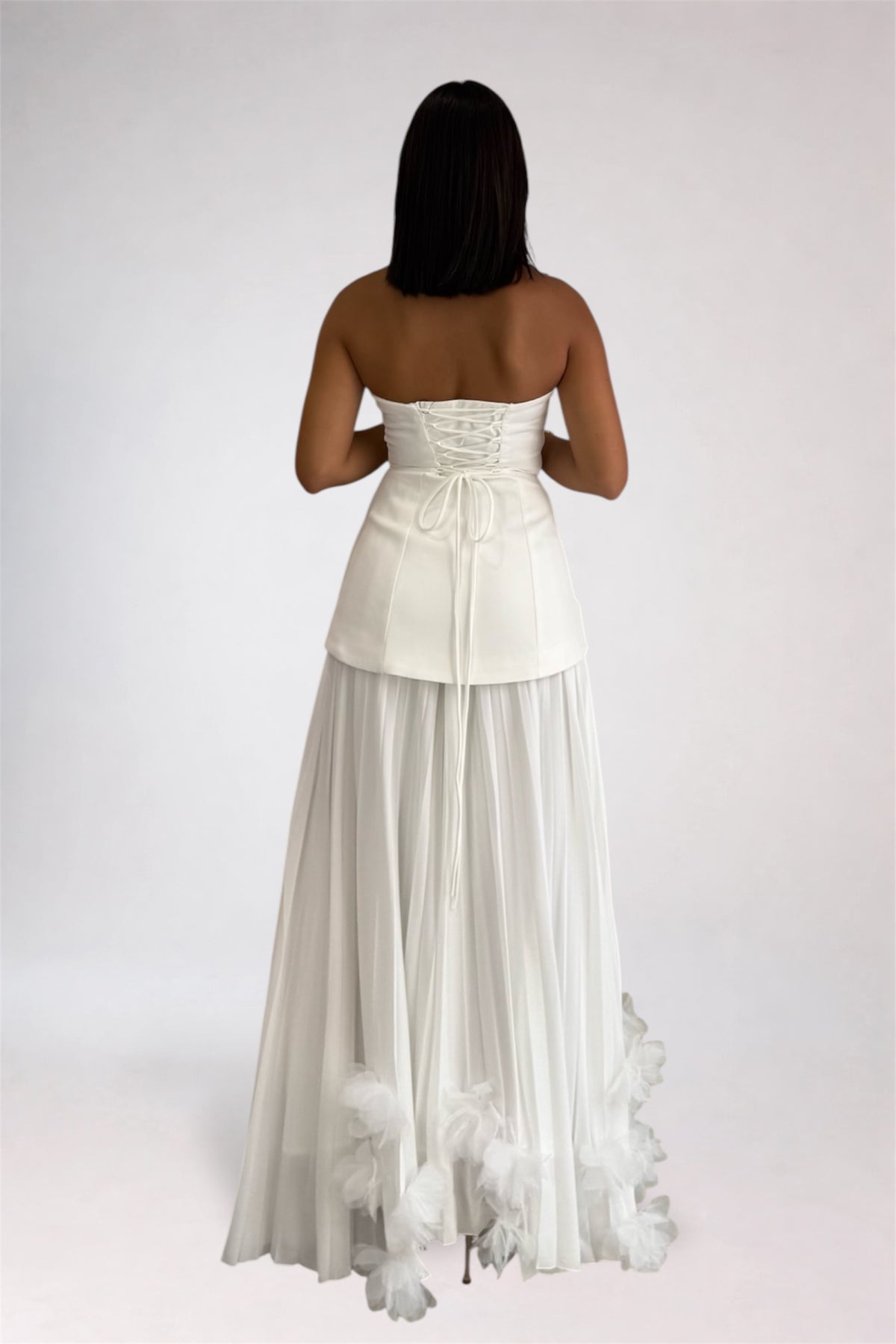 A woman viewed from the back wearing a strapless white formal gown with a structured corset-style waist, sheer long skirt, and feathered trim near the hem, against a plain light gray studio background.