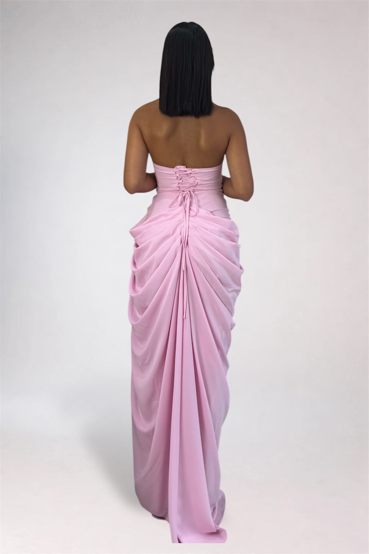 A woman viewed from behind wearing a strapless light pink maxi dress with a draped, gathered back design and a long train, photographed against a plain light gray studio background.