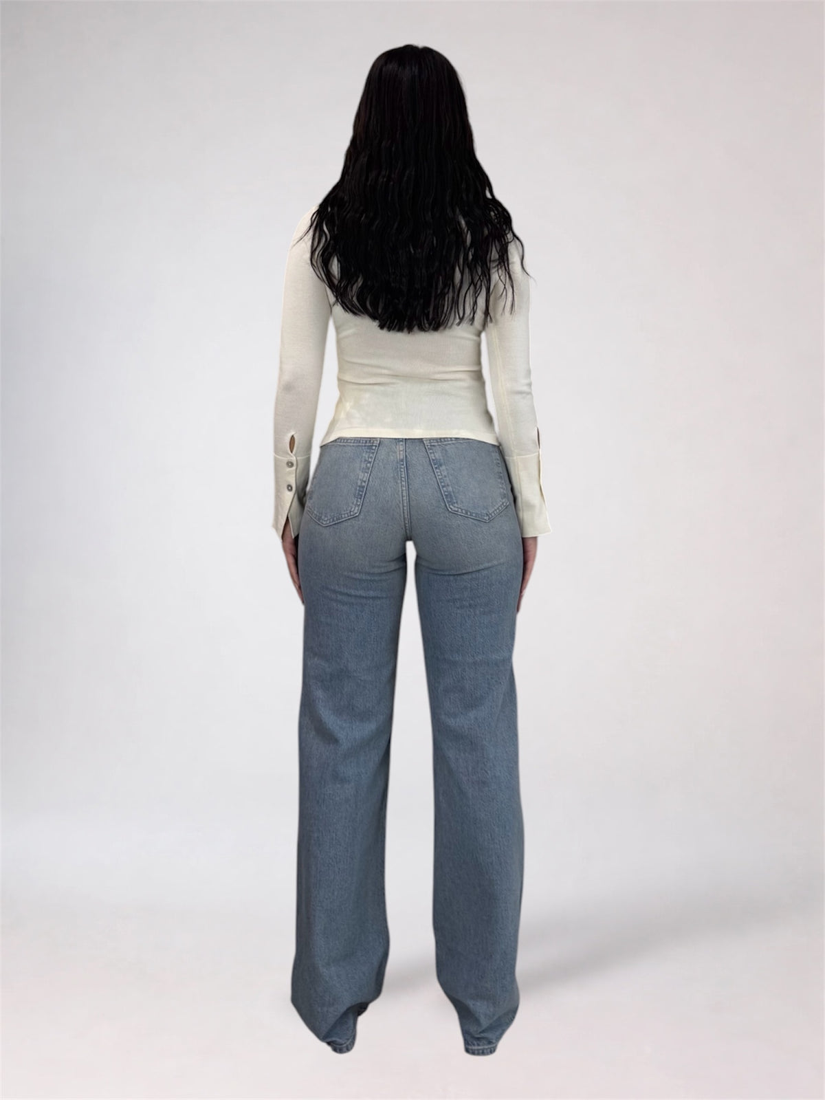 A woman viewed from behind wearing a fitted off-white long-sleeve top and light blue wide-leg jeans against a plain light gray studio background.