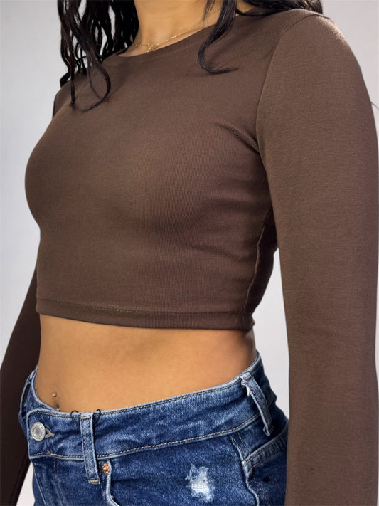A woman wearing a fitted, long-sleeve cropped top in a solid dark brown color, paired with blue jeans, shown from the chest down against a neutral gray background.
