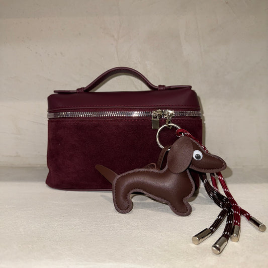 Brown dachshund-shaped keychain with plaid ribbon attached to a maroon handbag on a light gray surface.