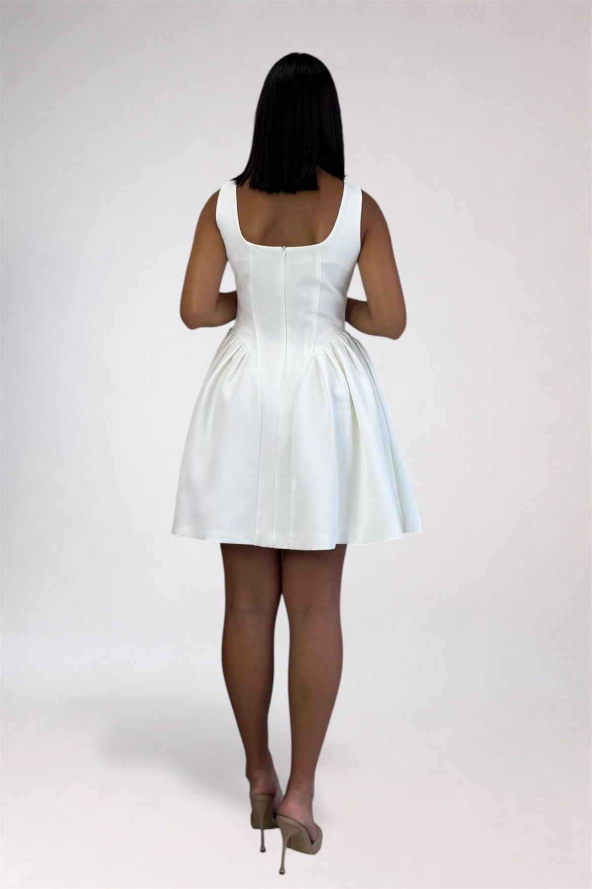 A woman viewed from behind wearing a white sleeveless mini dress with a structured back and gathered skirt, paired with beige high heels, against a plain light gray studio background.