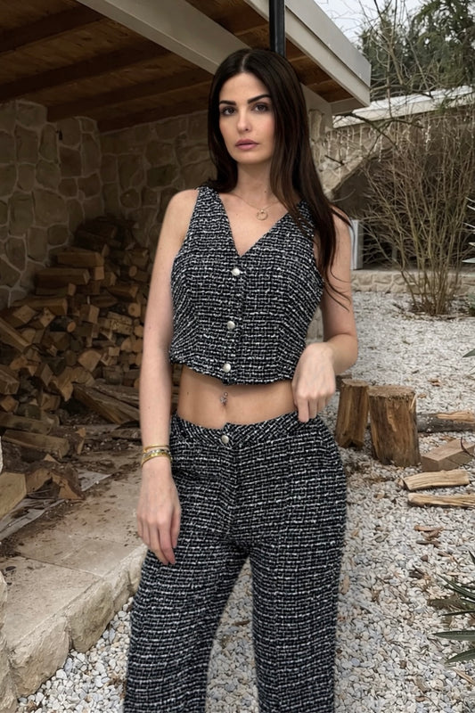 Woman wearing a black and white patterned outfit standing outdoors with wooden logs and a stone wall in the background.