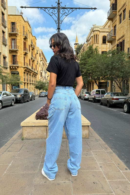 Woman in black t-shirt and light blue jeans standing on a street with buildings and cars in the background.