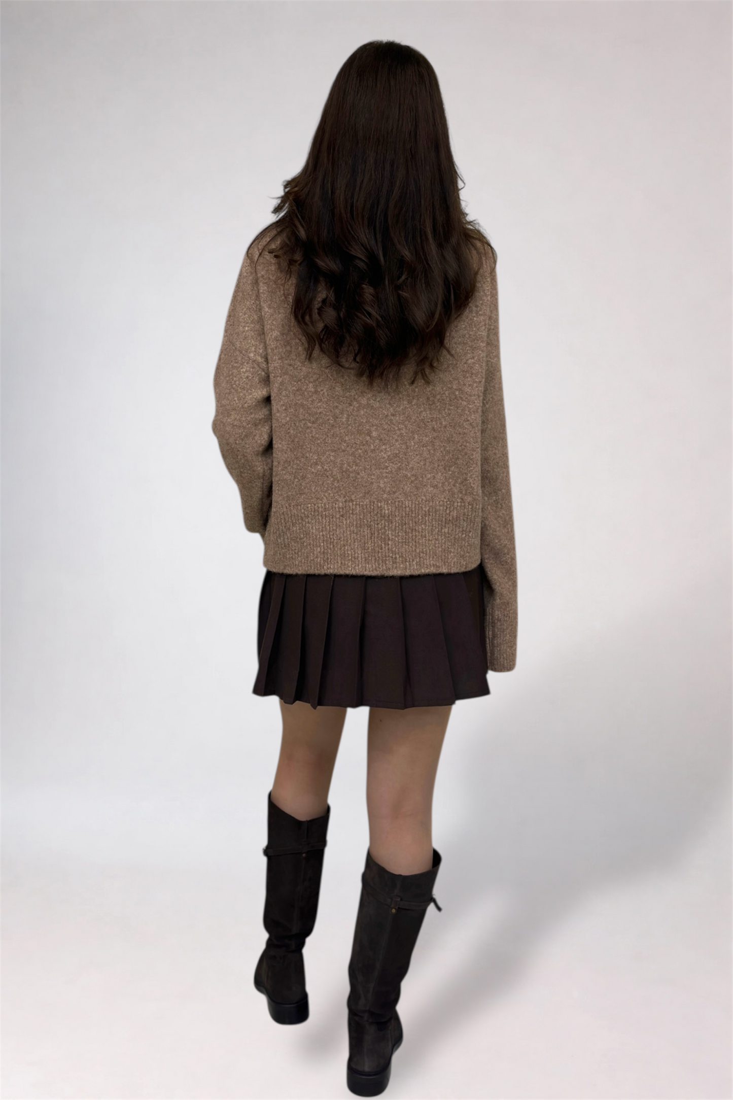 Person wearing a beige sweater, black pleated skirt, and black boots on a white background
