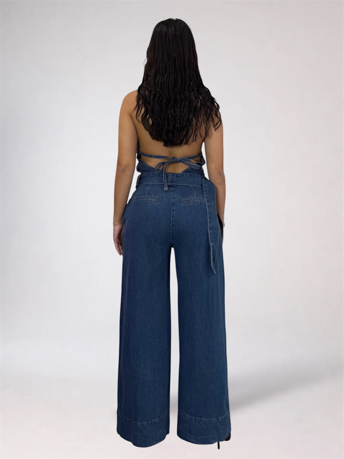 A woman viewed from the back wearing a dark blue denim jumpsuit with wide legs and a tie-back waist, against a plain light gray studio background.