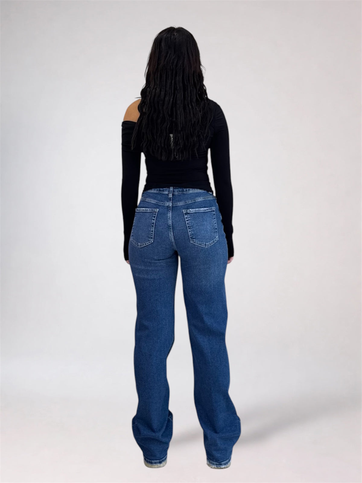 A woman viewed from the back wearing a black off-the-shoulder long-sleeve top and blue straight-leg jeans against a light gray studio background.