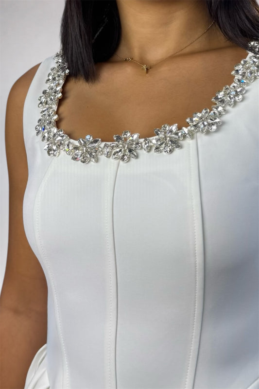 Close-up of a woman wearing a white sleeveless dress with a wide, embellished scoop neckline featuring large clear rhinestone flowers and silver-toned metal settings.