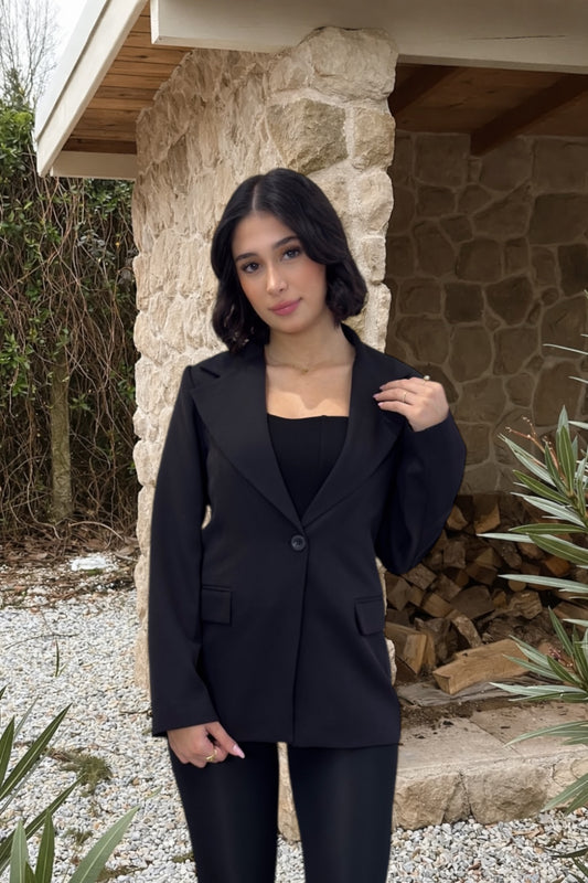 Woman in a black suit standing outdoors near a stone wall and wooden logs.