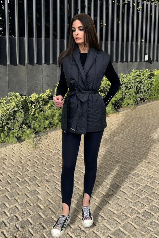 Woman wearing a black vest over a black top and leggings, standing on a paved path with greenery in the background.
