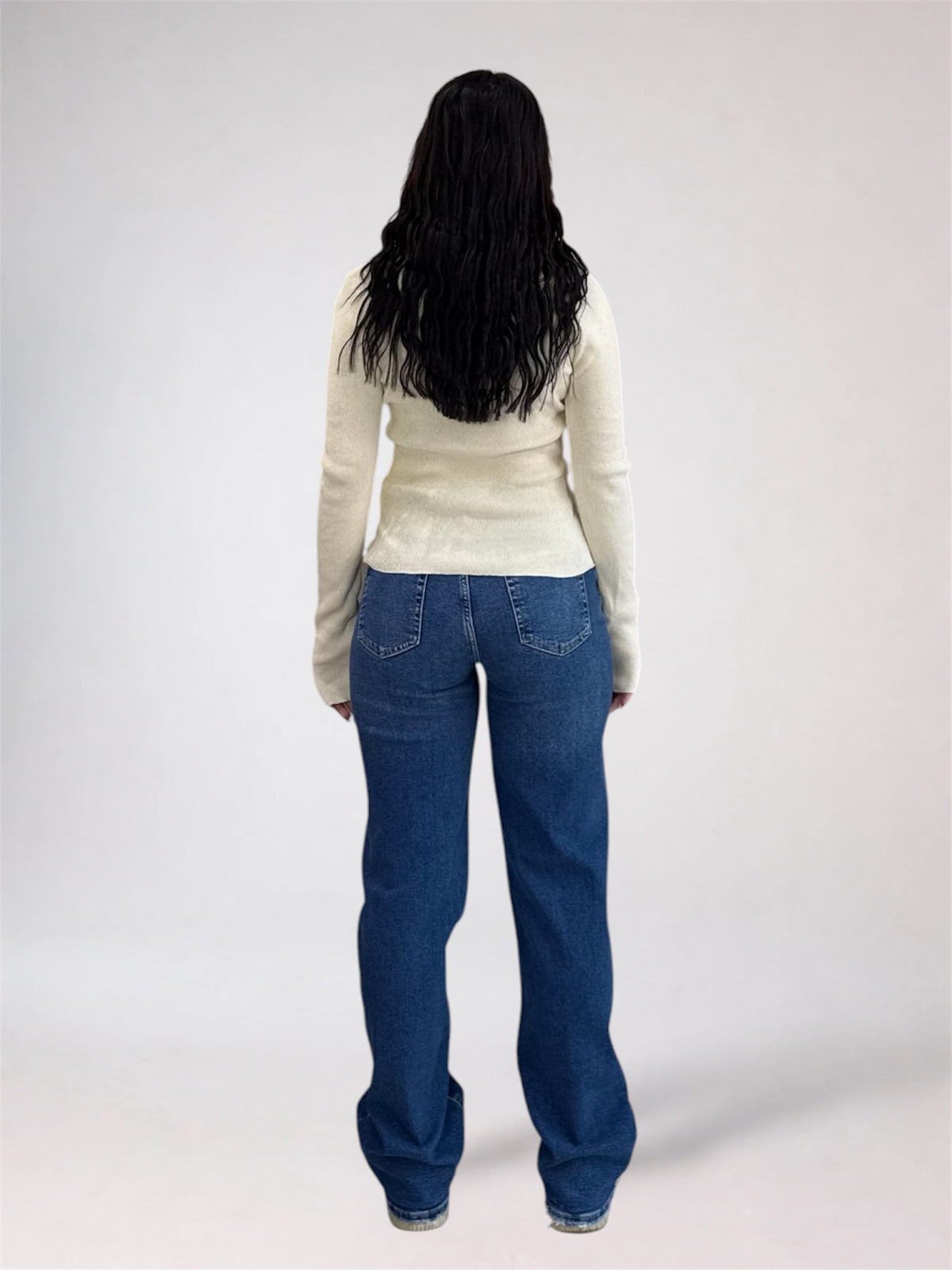 A woman viewed from behind wearing a cream-colored long-sleeve knit top and blue jeans, standing against a light gray studio background.