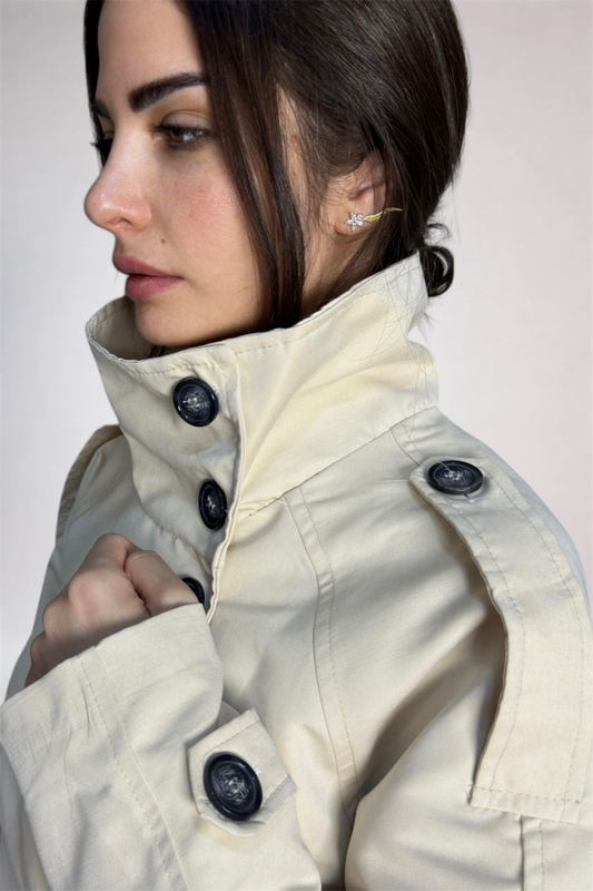 Woman wearing a beige trench coat with black buttons on a plain background