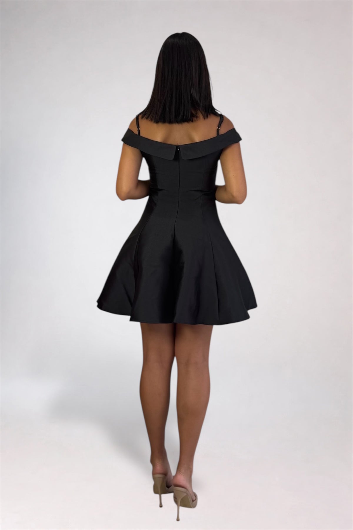 A woman viewed from behind wearing a short off-the-shoulder black dress with a flared skirt, paired with beige high heels, against a light gray studio background.