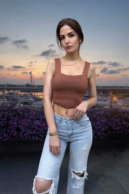 Woman in a brown top and light blue jeans standing on a rooftop with a cityscape and sunset in the background.