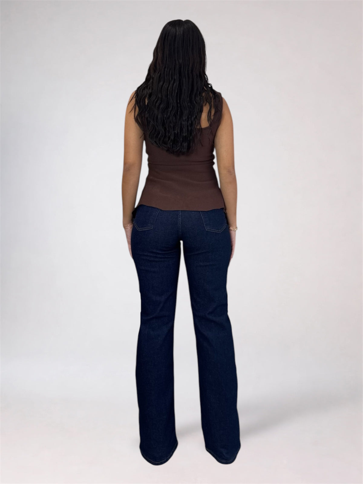 A woman viewed from behind wearing a brown sleeveless top and dark blue flared jeans against a light gray studio background.