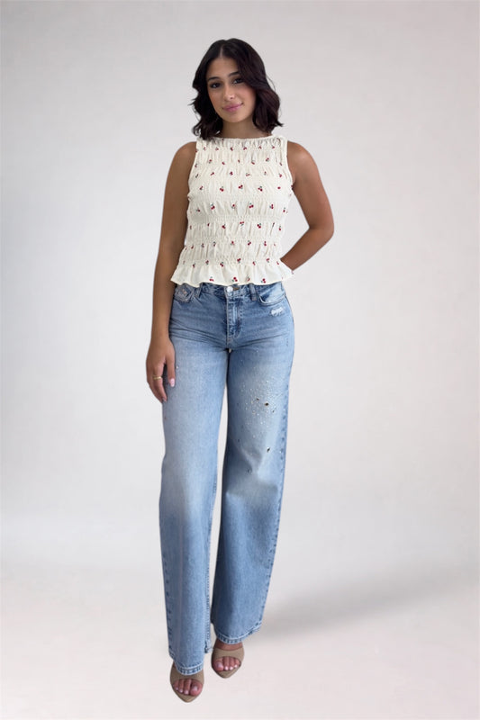Woman wearing a white sleeveless top with floral patterns and blue jeans on a plain background
