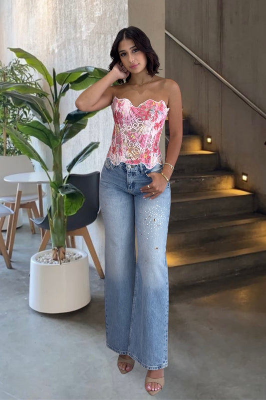 Woman in floral top and denim jeans standing indoors with a plant and staircase in the background