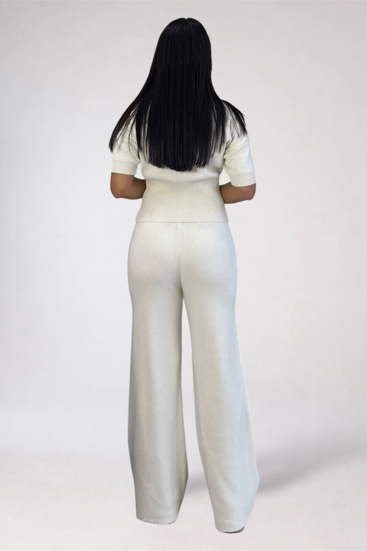 A woman viewed from behind wearing a matching cream-colored short-sleeve top and wide-leg trousers set, standing against a plain light gray studio background.