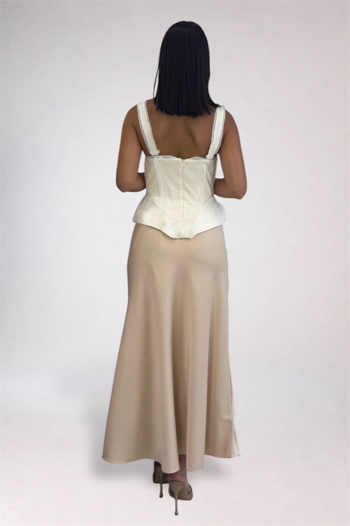 A woman shown from the back wearing a sleeveless cream corset-style top and a long beige skirt against a plain light gray studio background.