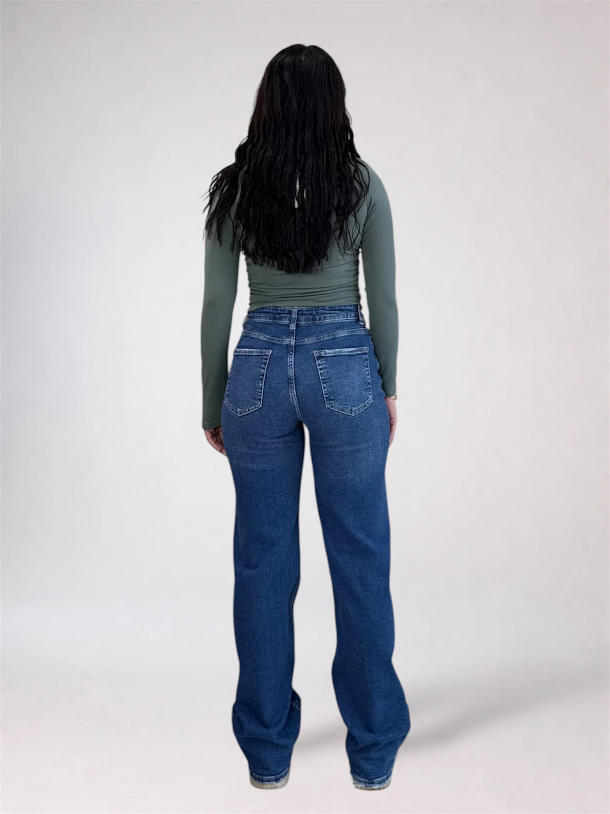 A woman viewed from behind wearing a sage green long-sleeve top and blue wide-leg jeans against a plain light gray studio background.