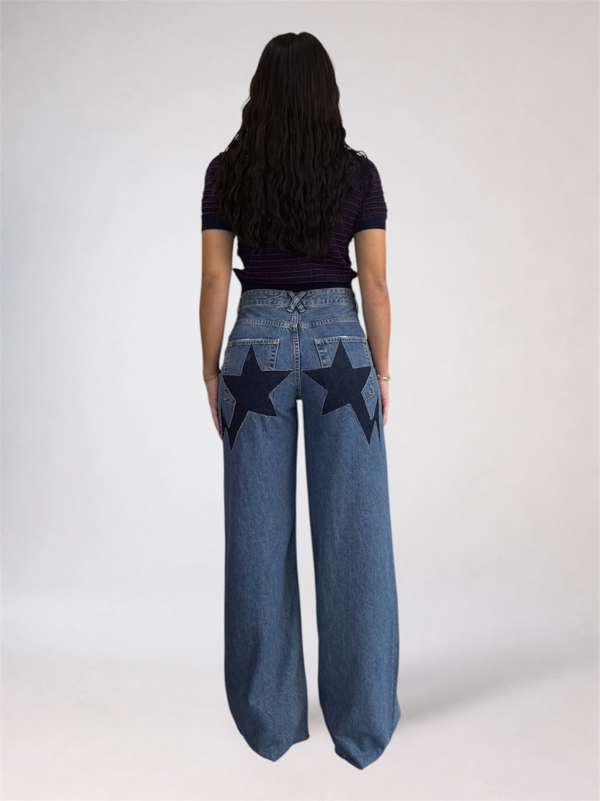 A model viewed from the back wearing high-waisted wide-leg blue jeans with large dark star patches on the back pockets, paired with a dark striped short-sleeve top, against a light gray studio background.