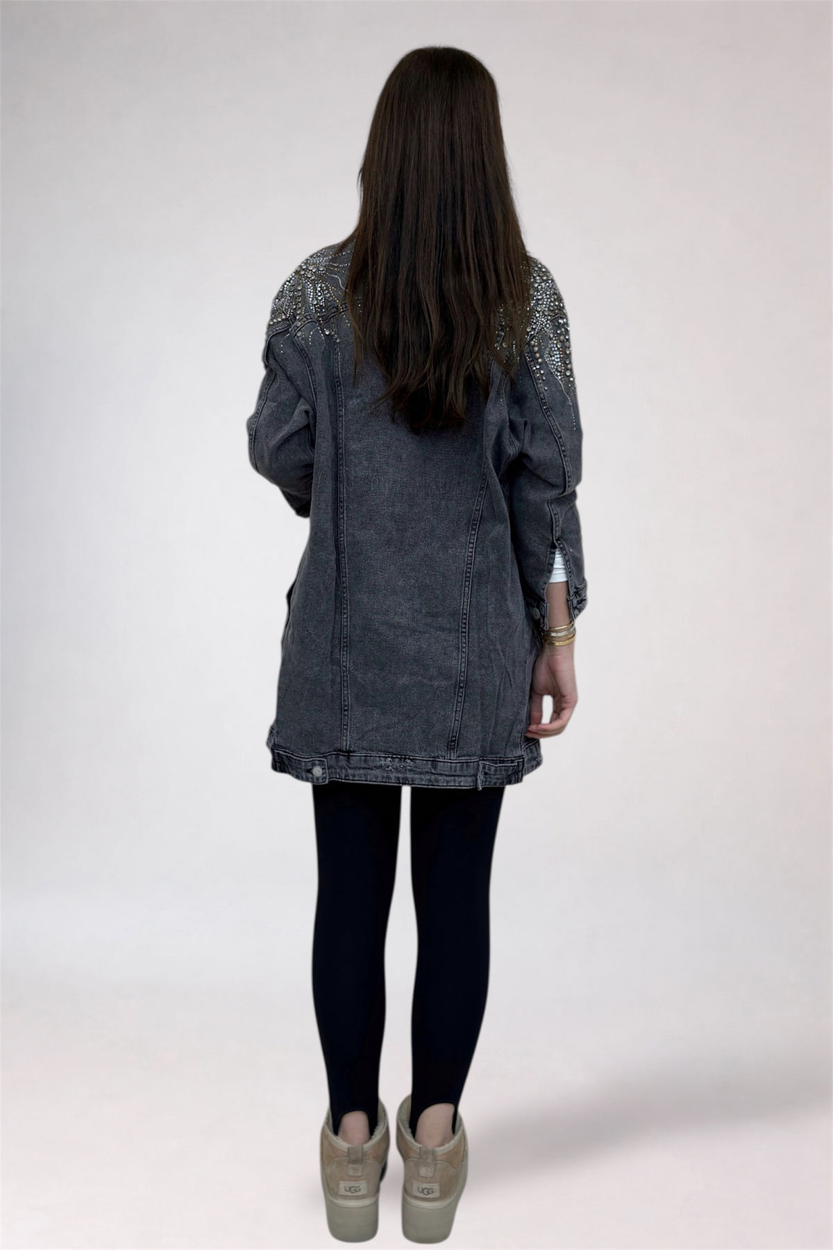 Person wearing a dark denim jacket with embellishments on a white background