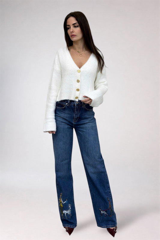 Woman wearing a white cardigan and blue jeans with embroidery on a plain background