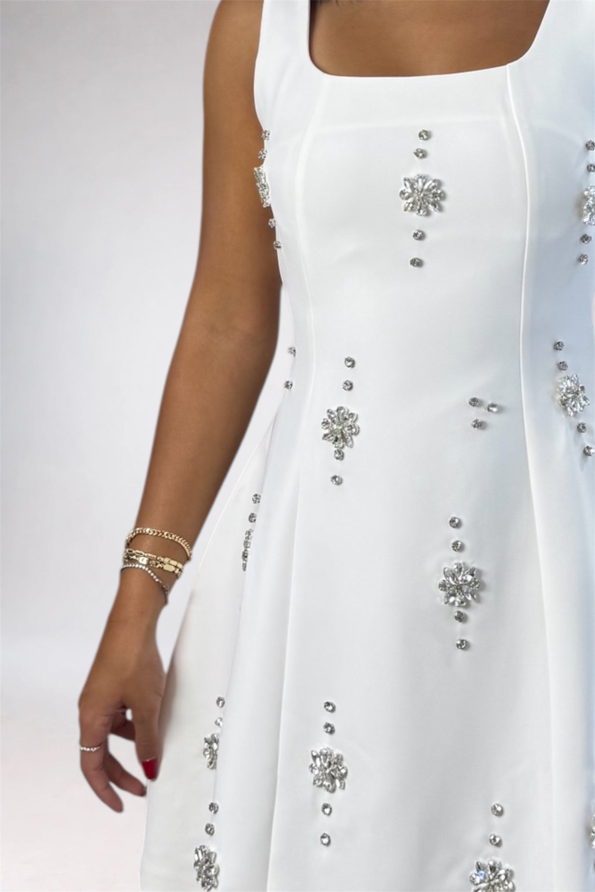 A close-up of a white sleeveless dress with silver rhinestone embellishments, worn by a model whose arm and hand are visible against a light gray studio background.
