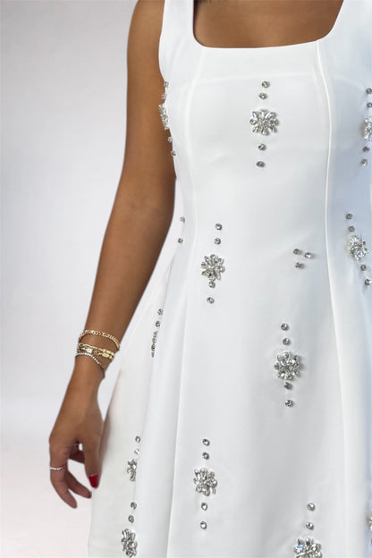 A close-up of a white sleeveless dress with silver rhinestone embellishments, worn by a model whose arm and hand are visible against a light gray studio background.