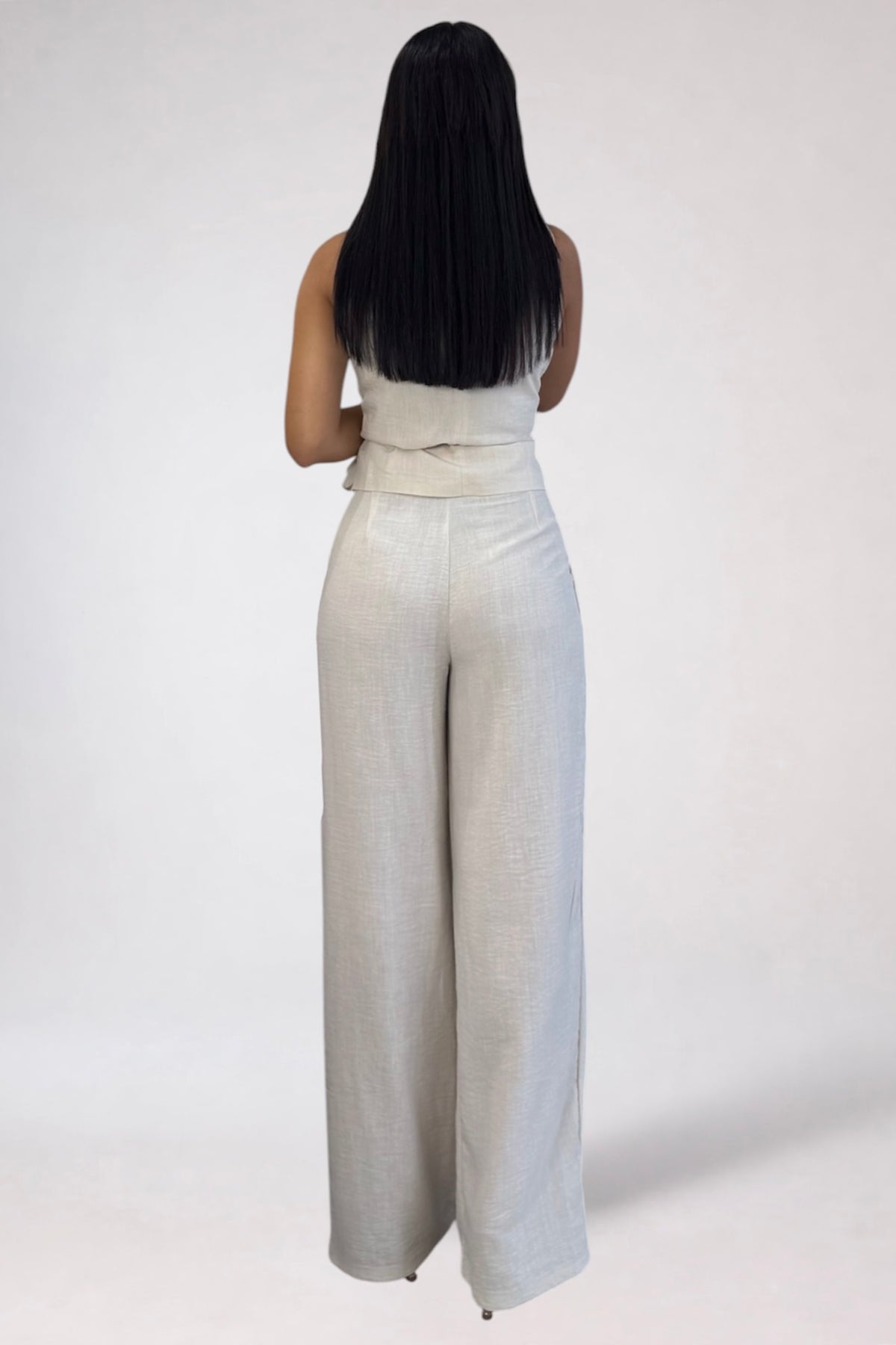 A woman viewed from behind wearing a light beige sleeveless jumpsuit with wide legs and a tied waist, standing against a plain light gray studio background.