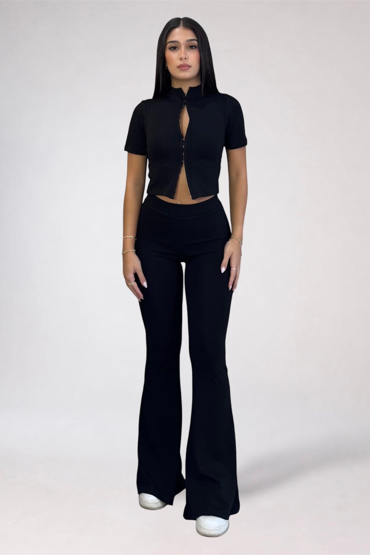 A woman modeling a black short-sleeve crop top with a front zipper and keyhole cutout, paired with matching black flared pants, against a plain light gray studio background.