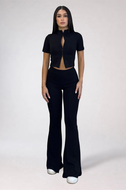 A woman modeling a black short-sleeve crop top with a front zipper and keyhole cutout, paired with matching black flared pants, against a plain light gray studio background.
