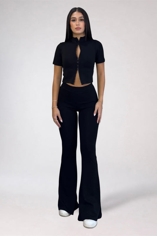 A woman modeling a black short-sleeve crop top with a front zipper and keyhole cutout, paired with matching black flared pants, against a plain light gray studio background.