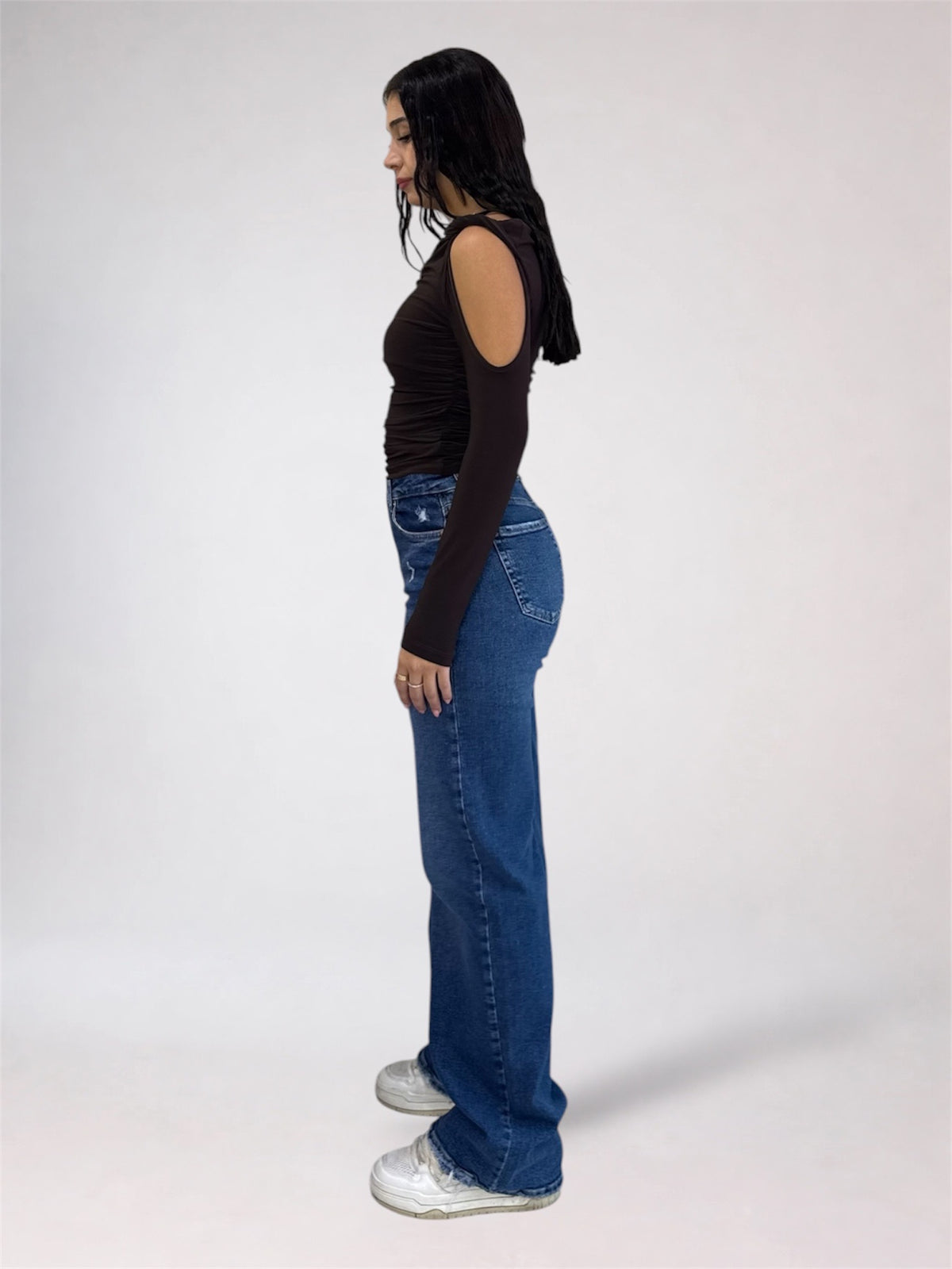 A woman standing in profile wearing a dark brown cold-shoulder long-sleeve top, blue wide-leg jeans, and white sneakers against a light gray studio background.