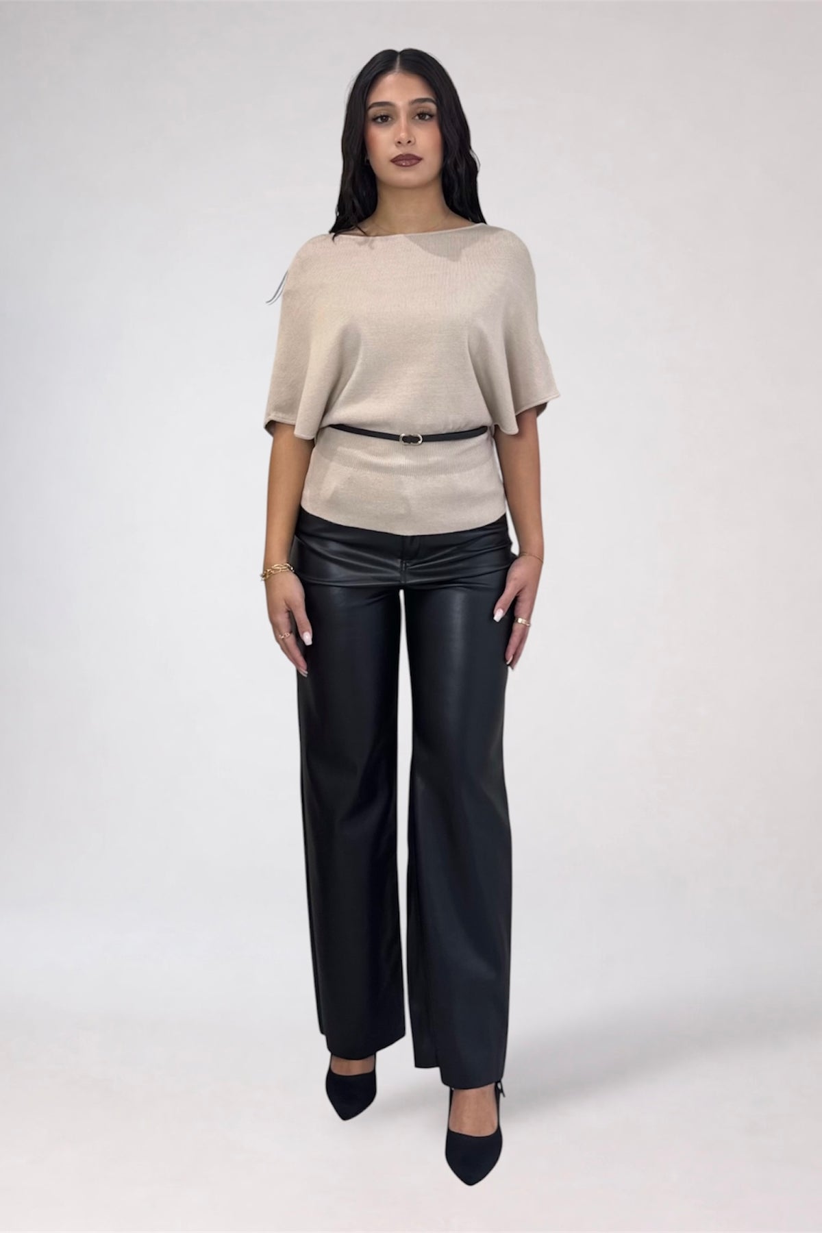 A woman modeling a light beige short-sleeve top with a thin black belt and black wide-leg pants, standing against a plain light gray studio background.