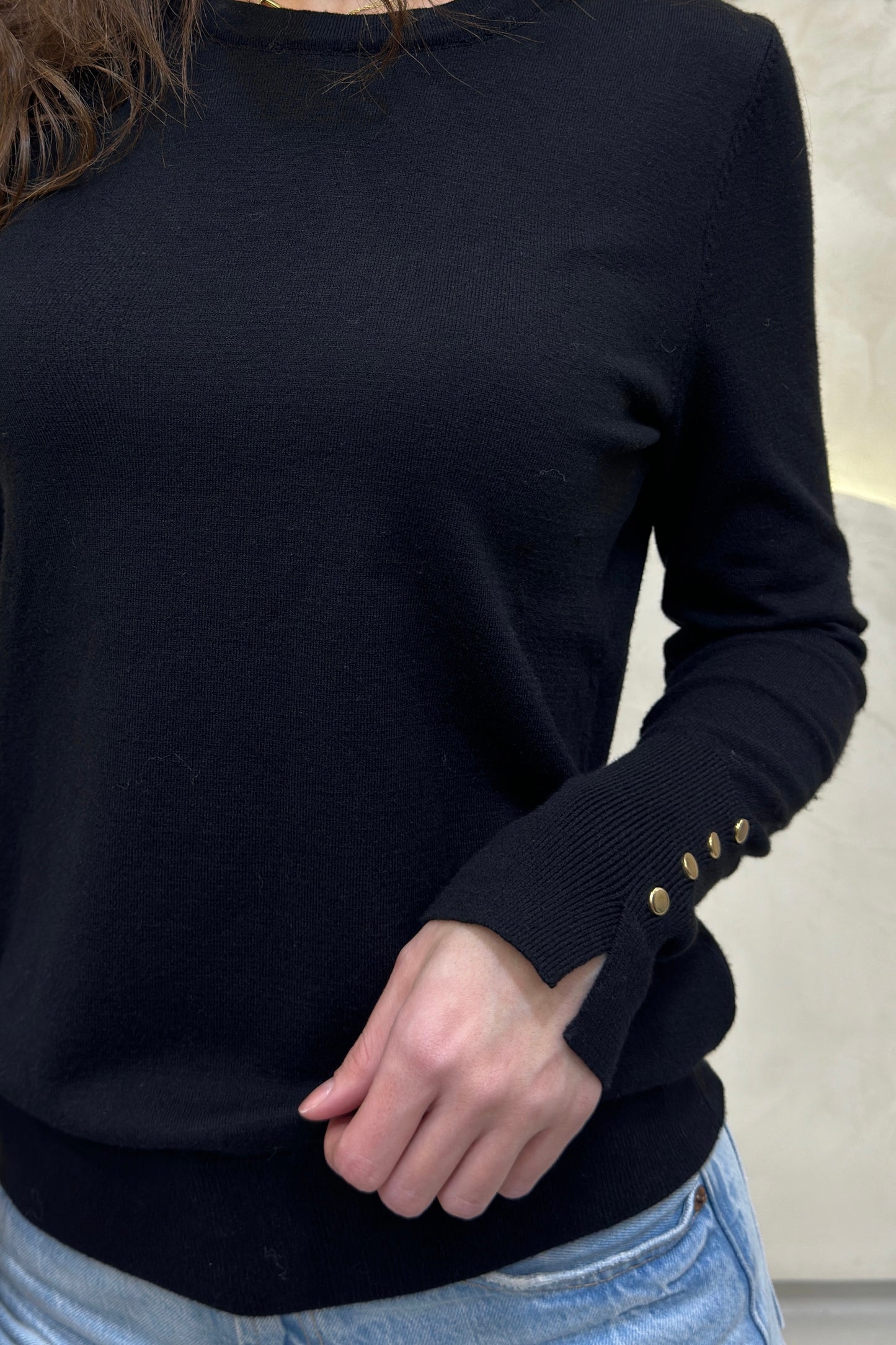 Black sweater with gold buttons on cuffs worn by a person, against a neutral background