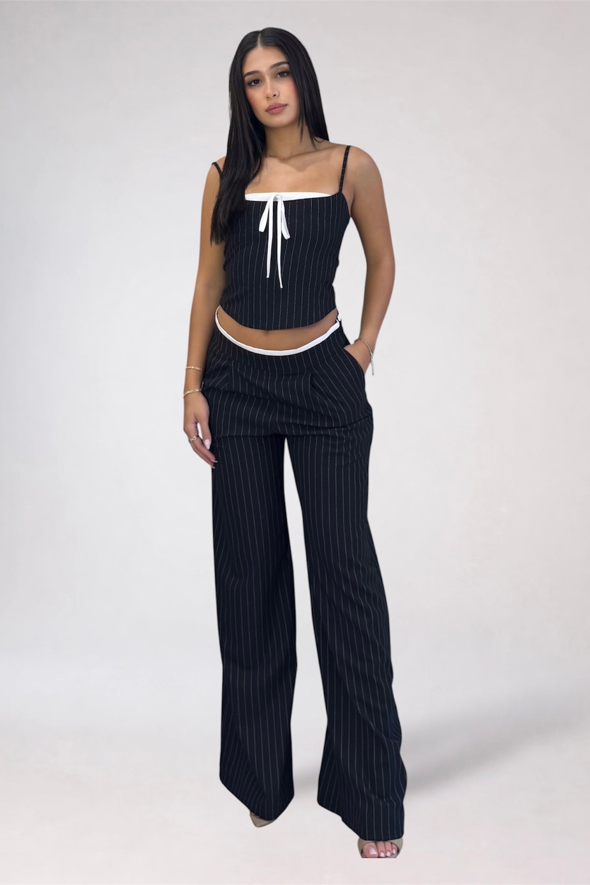 A woman modeling a matching two-piece pinstripe set consisting of a black camisole top with white trim and a white tie detail, paired with matching wide-leg pinstripe pants, against a light gray studio background.