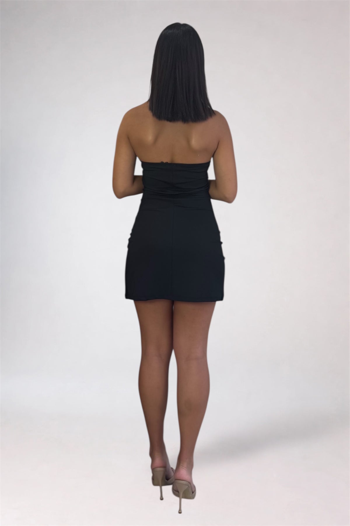 A woman viewed from behind wearing a strapless black mini dress and nude high heels against a plain light gray studio background.
