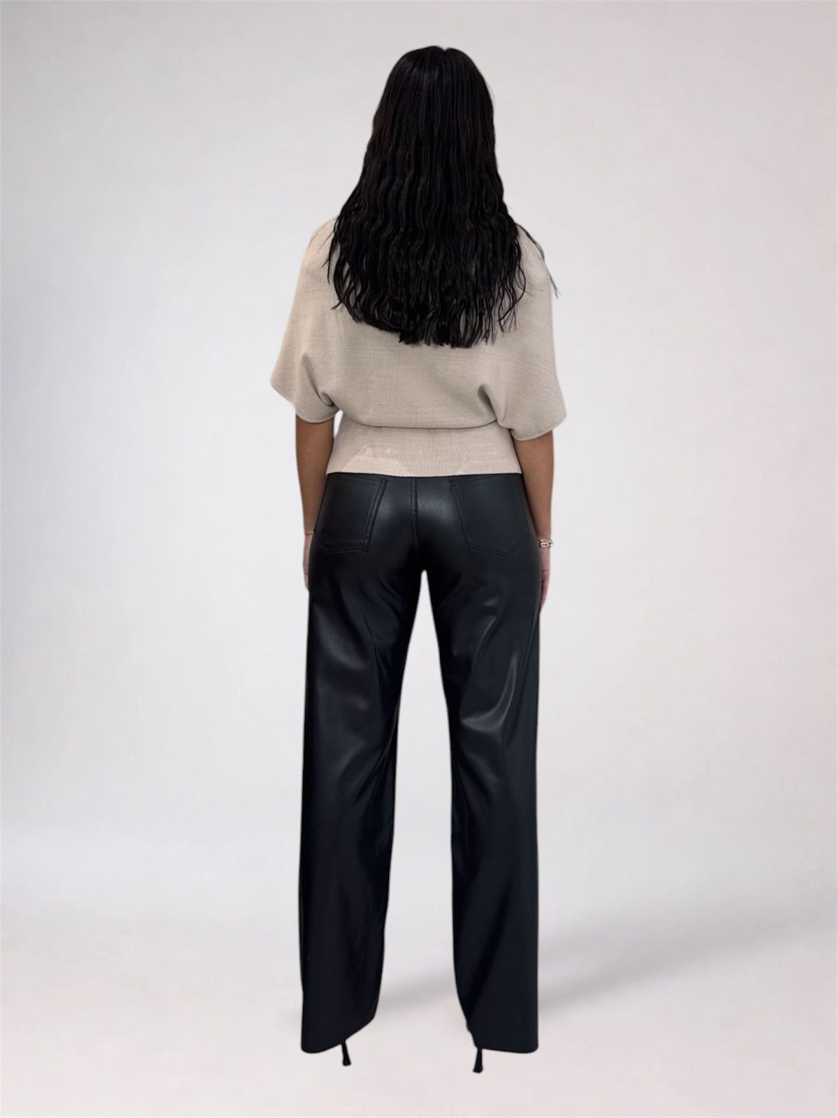 A model shown from the back wearing black wide-leg faux leather pants and a beige short-sleeve knit top, set against a plain light gray studio background.