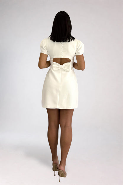 A woman viewed from behind wearing a short cream-colored dress with a cutout at the back and a bow detail, paired with beige high heels, against a light gray studio background.