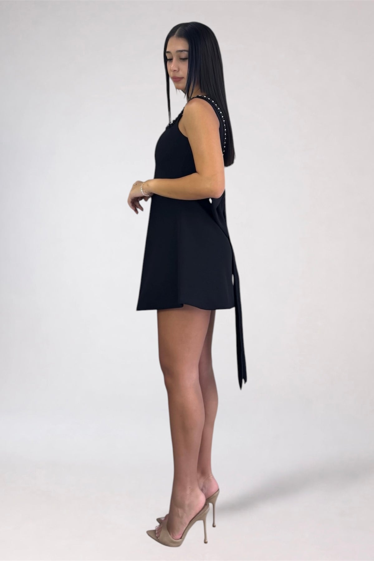 A woman modeling a black sleeveless mini dress with a side tie detail, paired with clear high-heeled sandals, photographed against a plain light gray studio background.