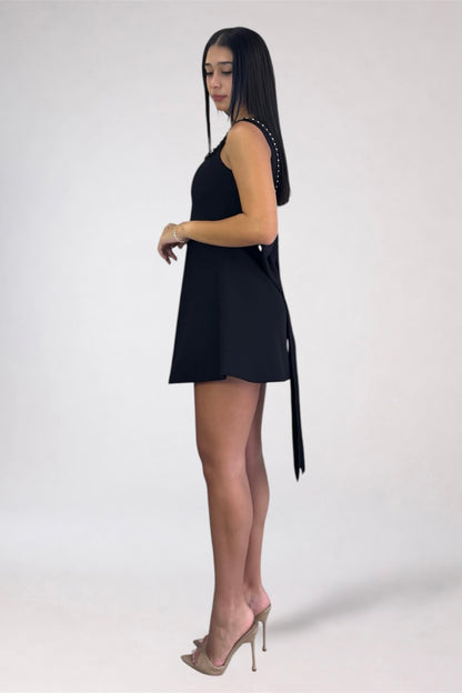 A woman modeling a black sleeveless mini dress with a side tie detail, paired with clear high-heeled sandals, photographed against a plain light gray studio background.