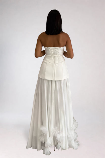 A woman viewed from the back wearing a strapless white formal gown with a structured corset-style waist, sheer long skirt, and feathered trim near the hem, against a plain light gray studio background.