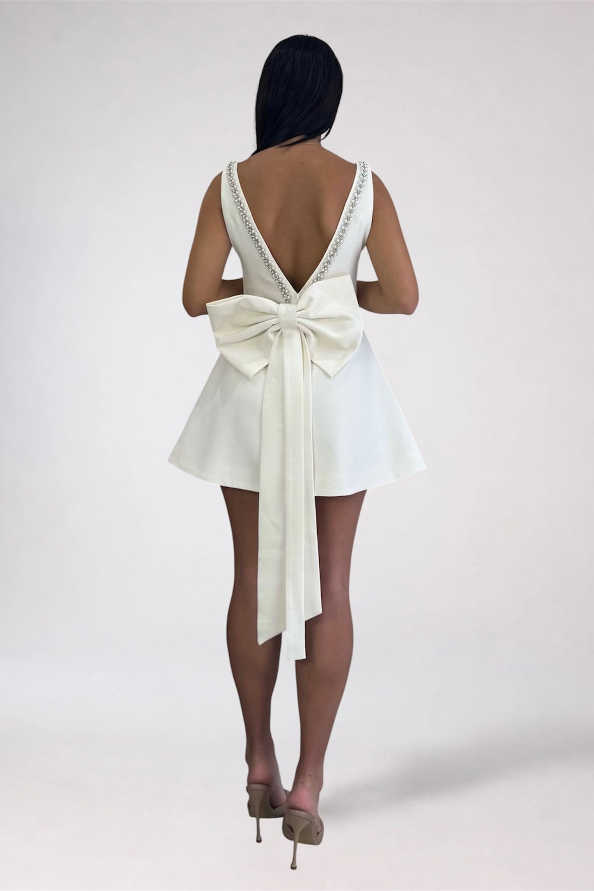 A woman shown from the back wearing a short ivory dress with a large bow at the back and a deep open back, photographed against a plain light studio background.