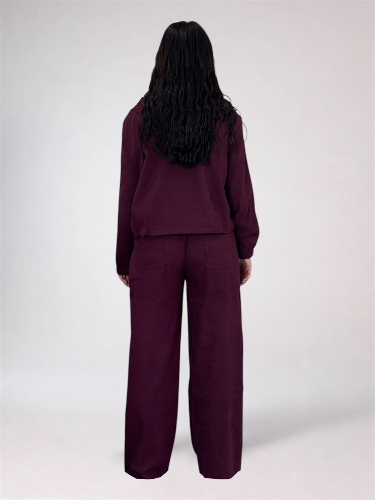 A model shown from the back wearing a matching deep plum purple two-piece set consisting of a cropped long-sleeve top and wide-leg trousers, set against a plain light gray studio background.