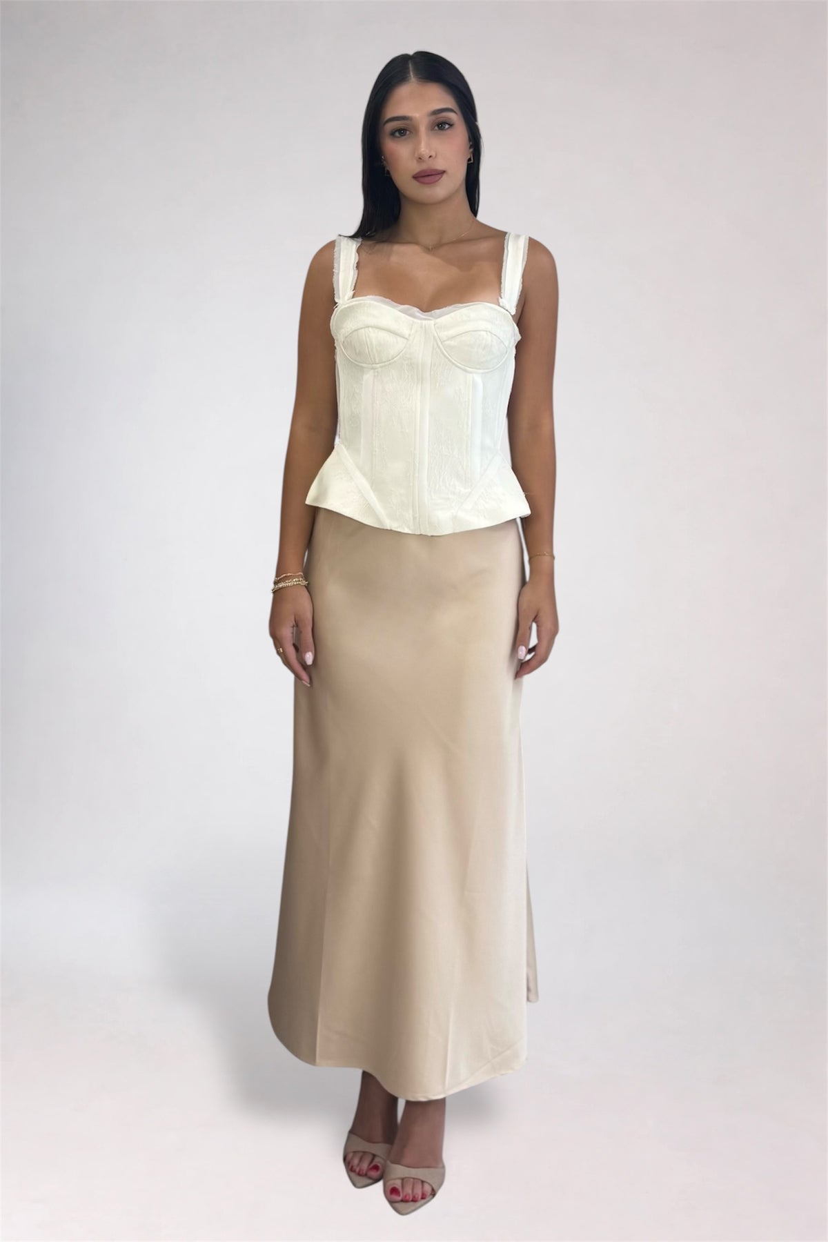 A woman modeling a two-piece outfit consisting of a cream corset-style camisole top and a matching beige maxi skirt, photographed against a plain light gray studio background.