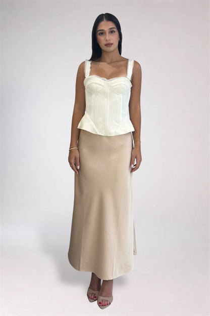A woman modeling a two-piece outfit consisting of a cream corset-style camisole top and a matching beige maxi skirt, photographed against a plain light gray studio background.