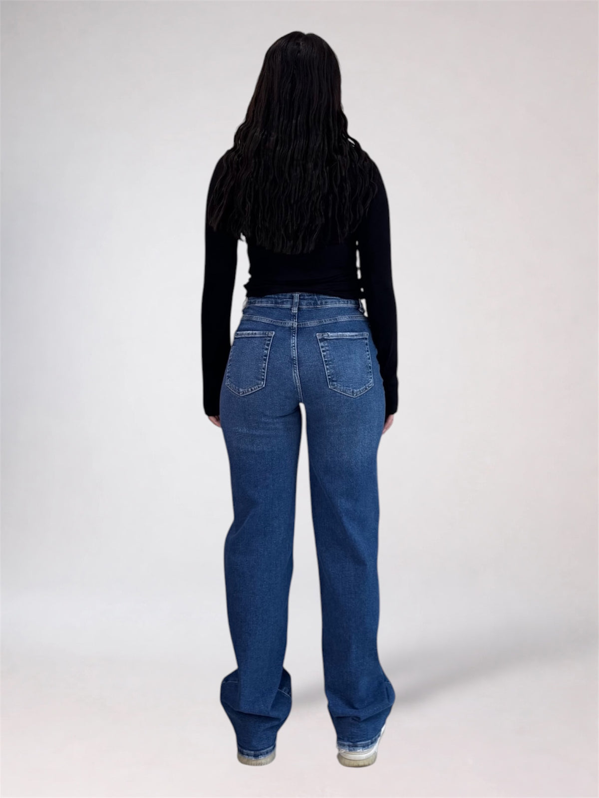 A person shown from the back wearing blue wide-leg jeans and a black long-sleeve top, standing against a plain light gray studio background.