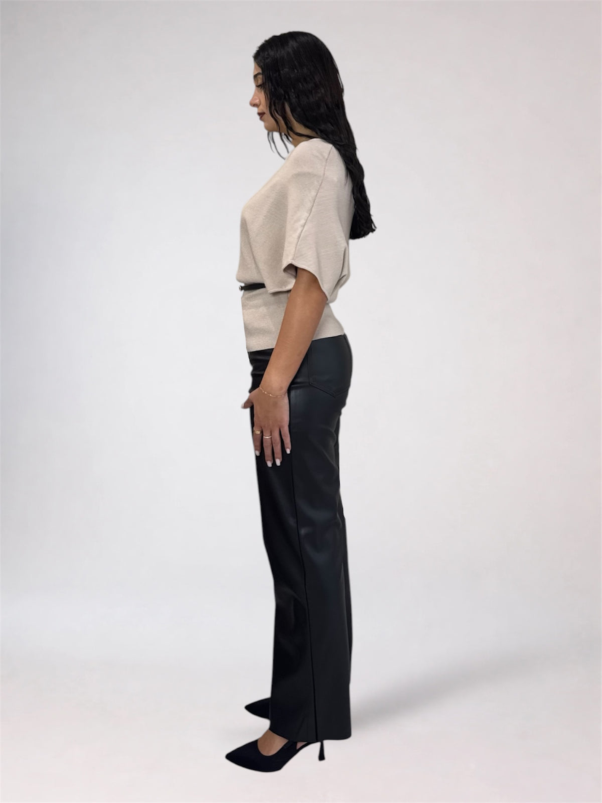 A woman shown in profile wearing a light beige short-sleeve top with a thin dark waistband detail and black wide-leg trousers, standing against a plain light gray studio background.